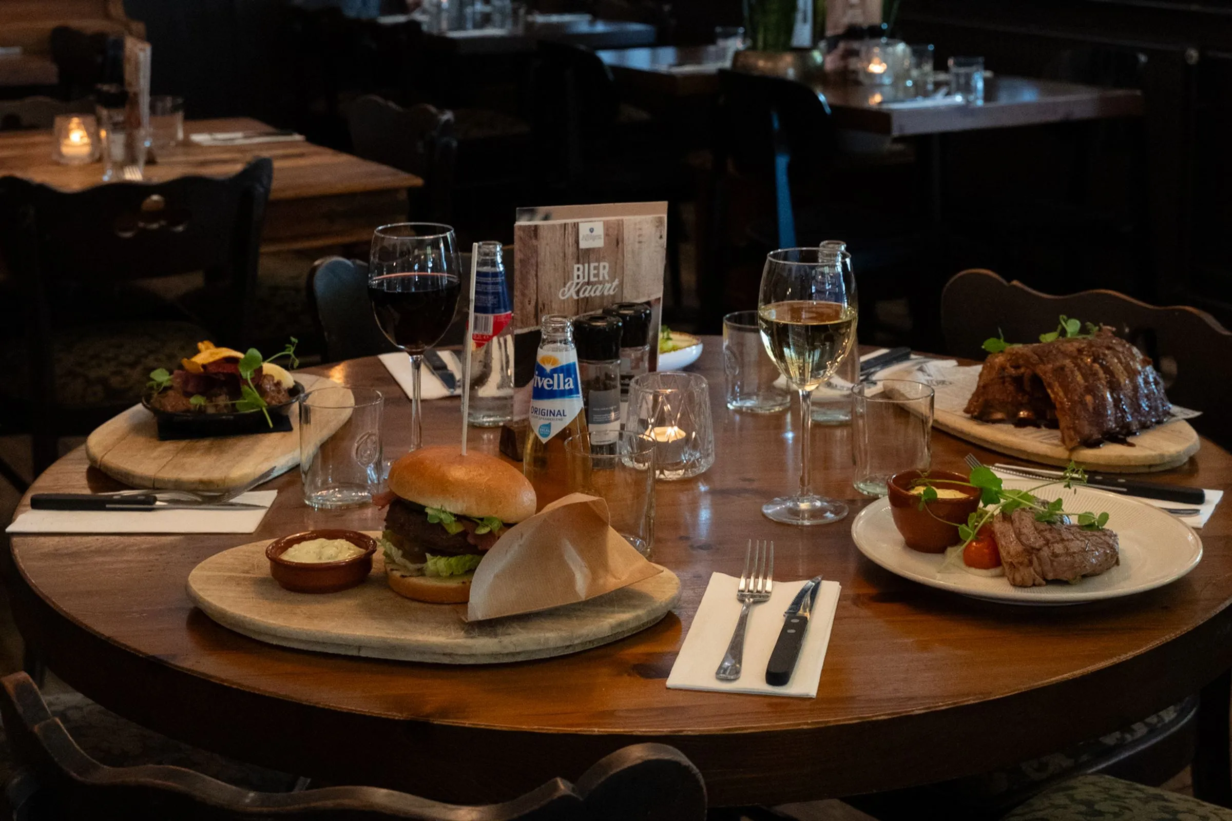 Een ronde houten tafel bij De Rosier met een burger, een steak, wijn en Belgisch bier onder een hanglamp